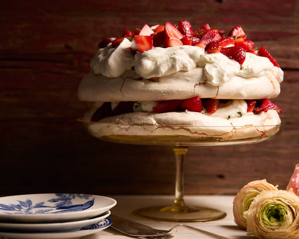 A strawberry pavlova dessert with layers of meringue and cream, garnished with fresh strawberries, on a gold pedestal plate beside a floral plate.