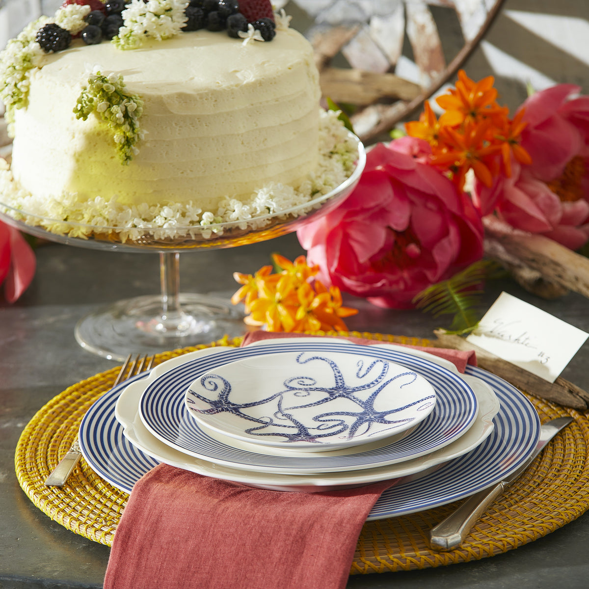A decorated cake on a stand next to a place setting with a red napkin under layered Starfish Small Plates by Caskata Artisanal Home on a yellow placemat. Bright orange and pink flowers surround the setup, adding a touch of elegance with heirloom-quality dinnerware.