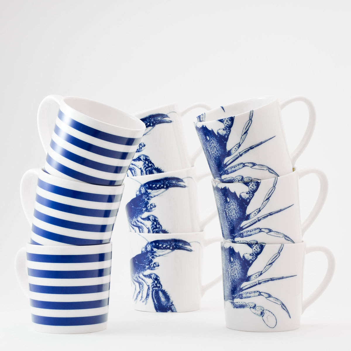 Two stacks of ceramic mugs are shown. One stack has blue and white stripes, while the other features blue crab illustrations on a white background. These Lobster Mugs by Caskata Artisanal Home are made in Sri Lanka.