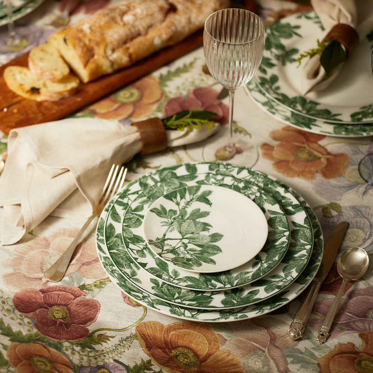 A table is set with green and white floral-patterned **Arbor Green Rimmed Dinner Plates** by **Caskata**, a wine glass, cutlery, and a cloth napkin. A bread loaf on a wooden board and a floral tablecloth are also visible.
