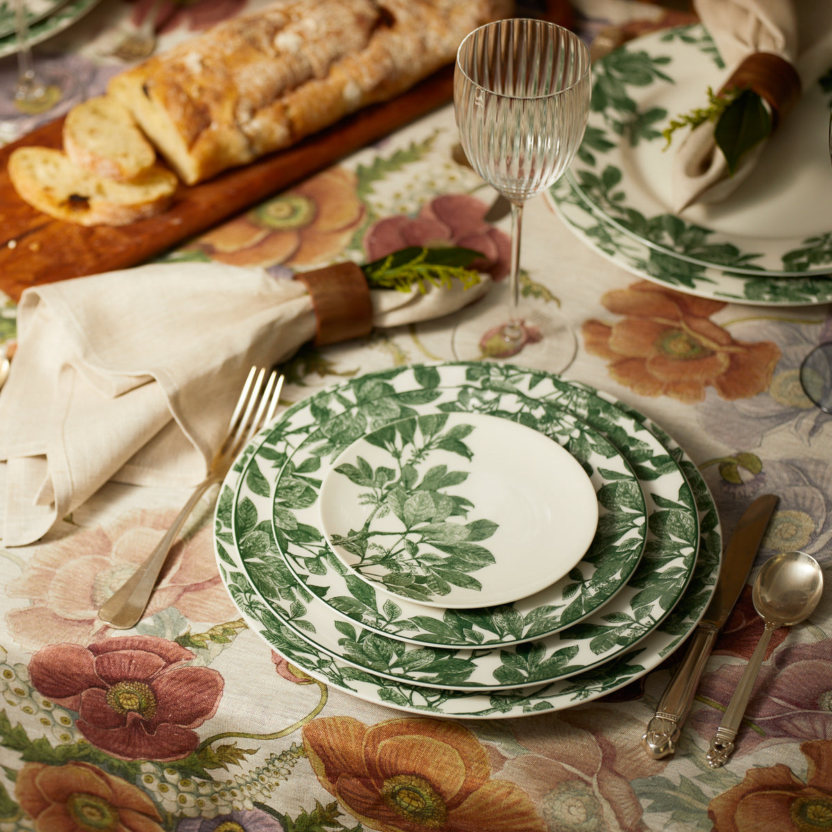 A table set with botanical dinnerware, featuring Caskata Arbor Green Small Plates, cutlery, wine glass, napkins with rings, and a loaf of bread on a matching floral tablecloth.