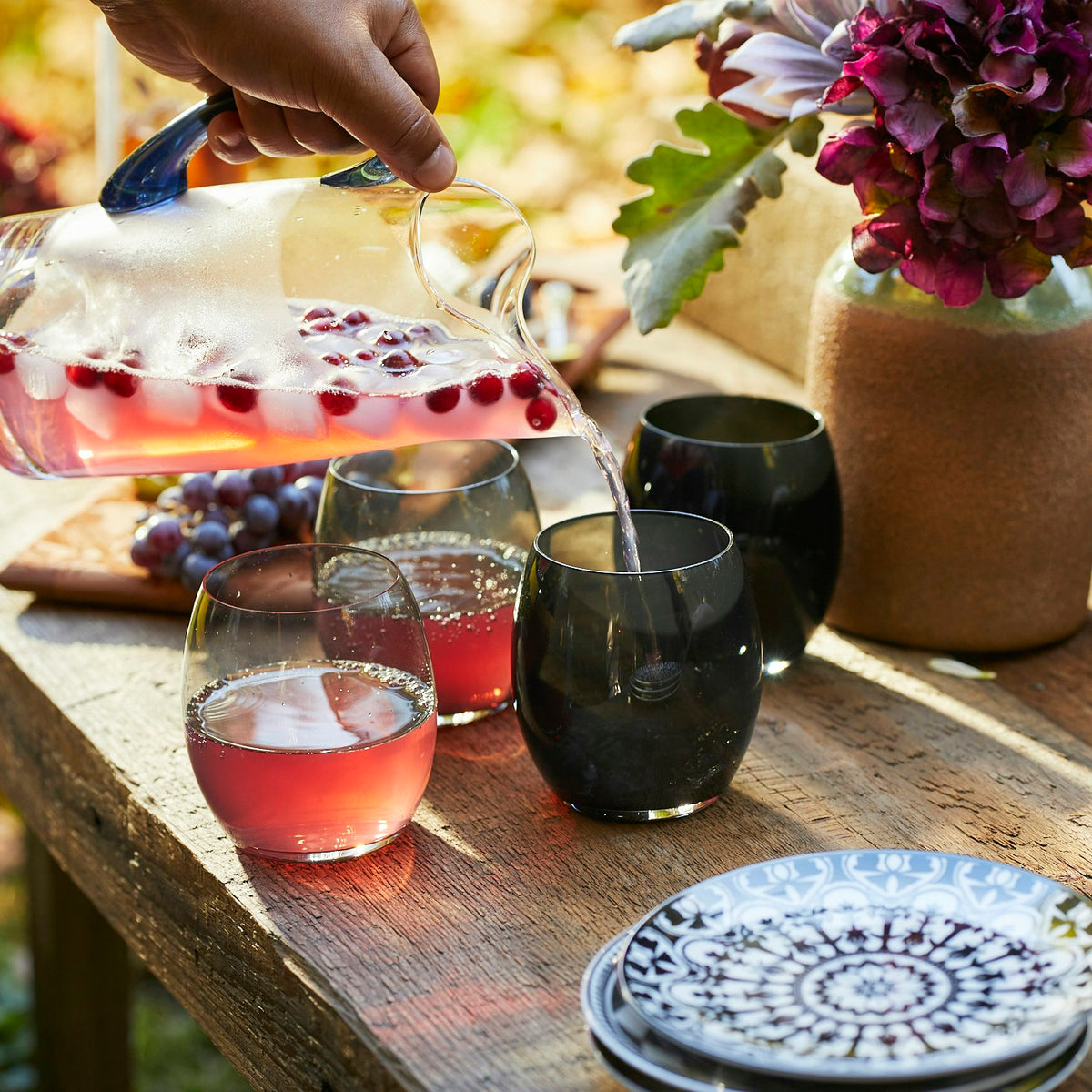 A person pouring colorful pomegranate juice from a Caskata Les Nuages Blue Handle Small Pitcher into lead-free crystal glasses on a table.
