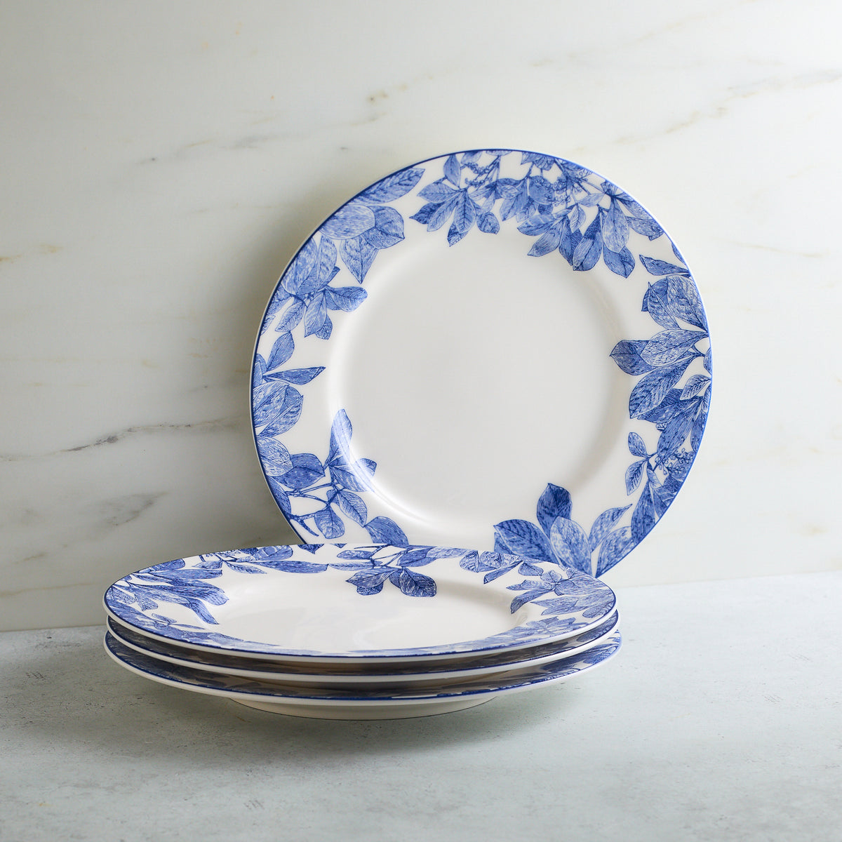 A stack of four Arbor Rimmed Salad Plates by Caskata Artisanal Home with blue leafy patterns on the edges, placed on a light gray surface against a marble background, evokes an heirloom feel.