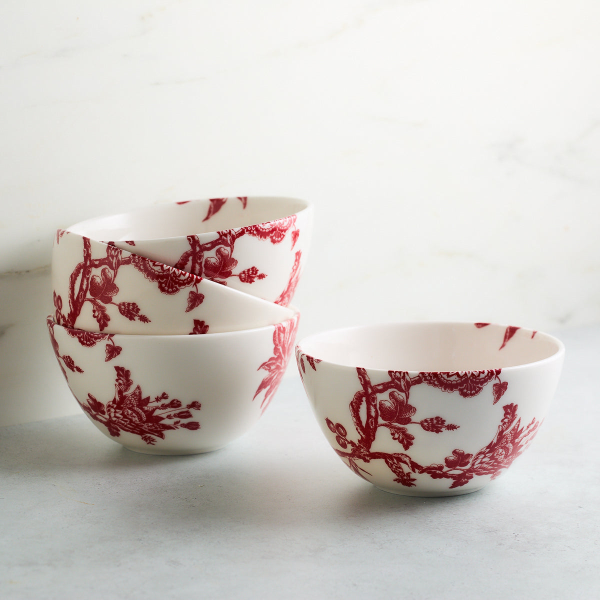 Three white porcelain bowls with red ornate designs are stacked, next to a fourth identical Arcadia Crimson Cereal Bowl by Caskata on a white and grey surface with a light background, showcasing the elegance of premium porcelain dinnerware inspired by the Williamsburg Foundation.