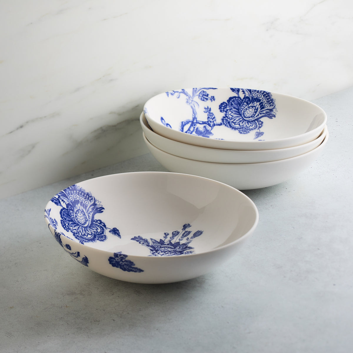 Four white ceramic bowls with blue floral patterns, crafted from premium Caskata Artisanal Home Arcadia Entrée Bowls. Three are stacked while one generous bowl is placed in front, all on a light gray surface with a white marble background. Inspired by the elegance of the Williamsburg Foundation.