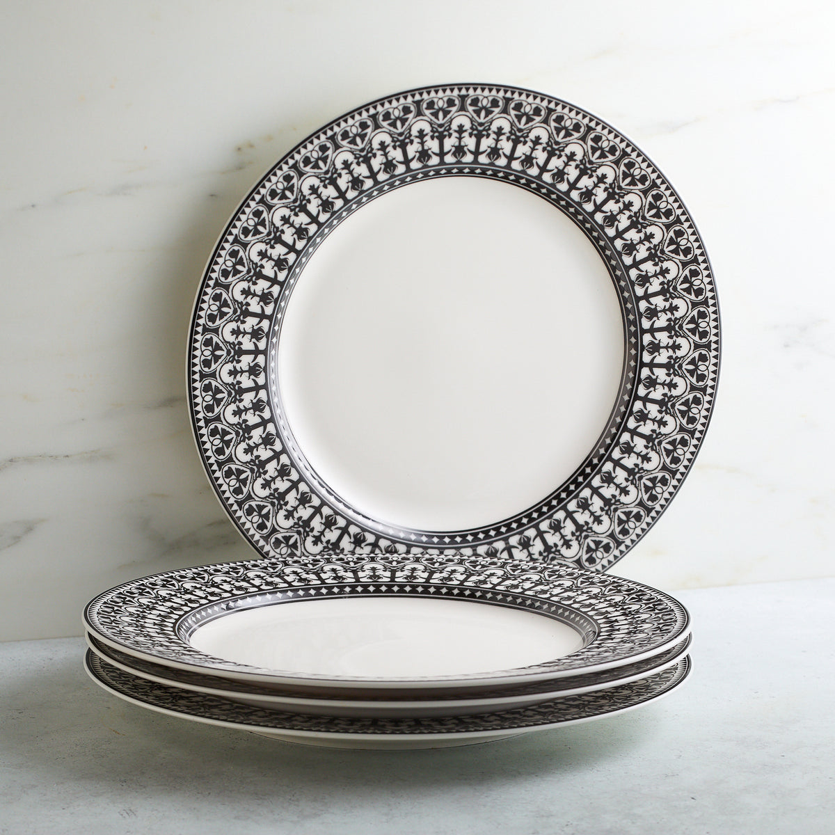 A stack of four white ceramic plates with intricate black, hand-decorated details around the edges, placed against a light background. This Casablanca Rimmed Dinner Plate by Caskata Artisanal Home exudes elegance and sophistication.