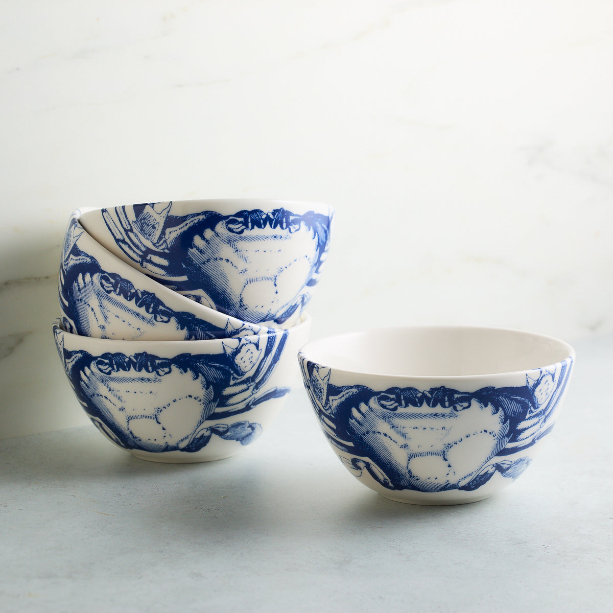 A stack of three blue and white high-fired porcelain bowls with a charming Crabs pattern on a marble surface, next to an additional matching Crab Cereal Bowl by Caskata to the right.