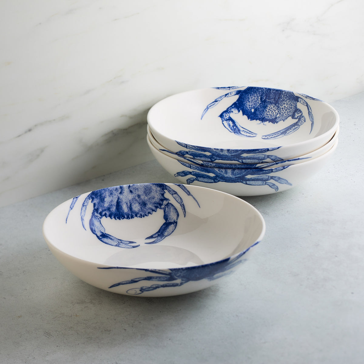 Four white ceramic bowls, each with a blue crab pattern, stacked next to a single identical bowl on a light grey surface. These Crab Entrée Bowls by Caskata Artisanal Home are high-fired porcelain and are dishwasher and microwave safe.