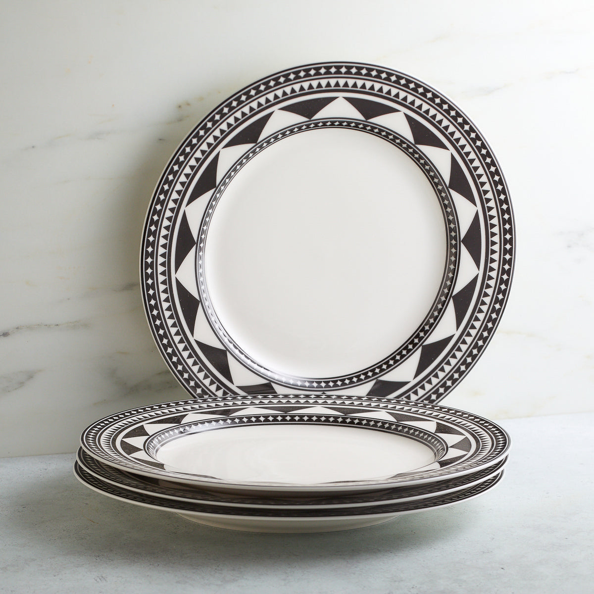 A stack of five Caskata Artisanal Home Fez Rimmed Dinner Plates with black graphic prints on the rims, placed on a light gray surface against a white marble background, showcases sophisticated dinnerware with subtle Moroccan patterns.