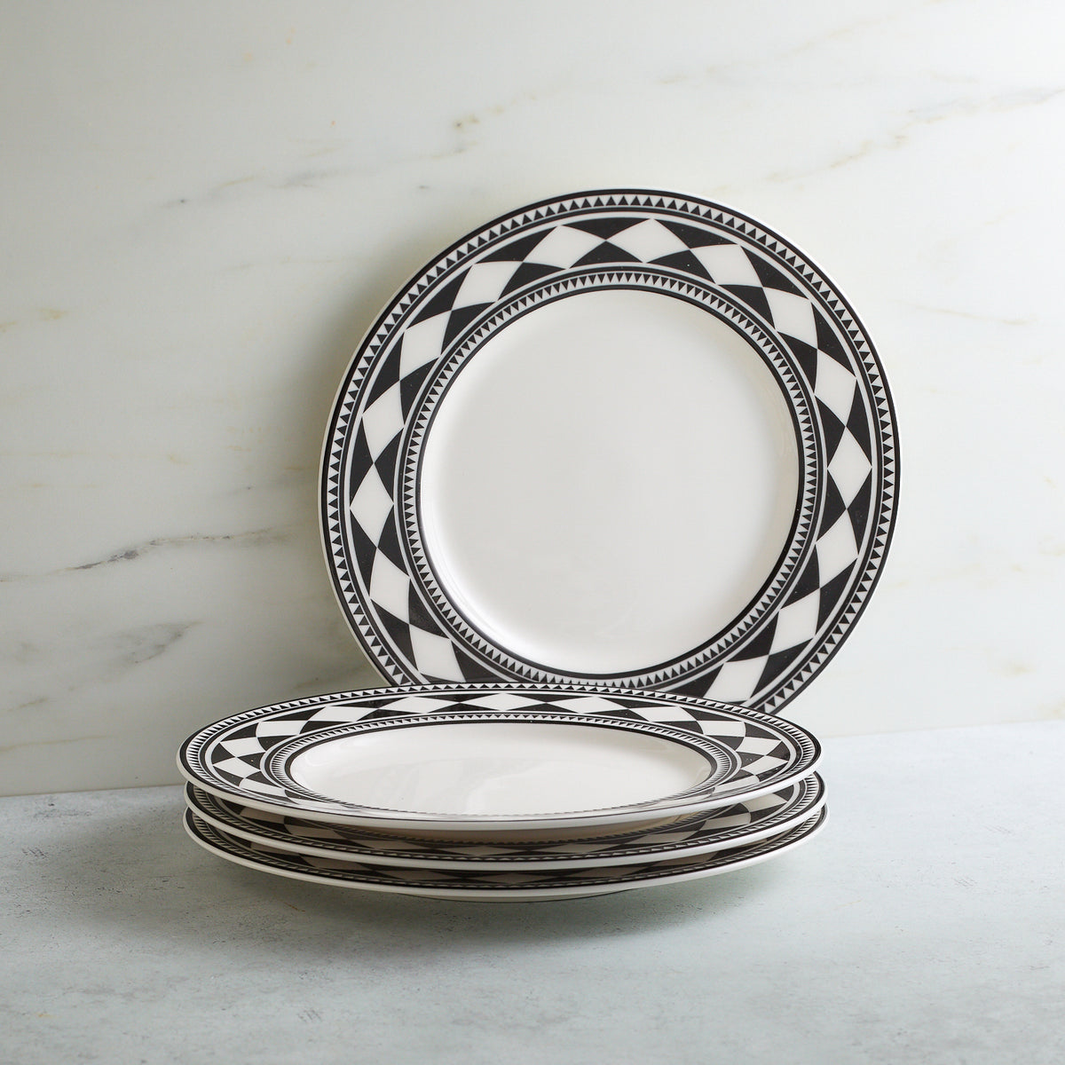 Stack of four Fez Rimmed Salad Plates by Caskata Artisanal Home with a sophisticated global feel and timeless shape, adorned with bold geometric patterns on the rim, positioned on a light-colored surface in front of a white marble background.