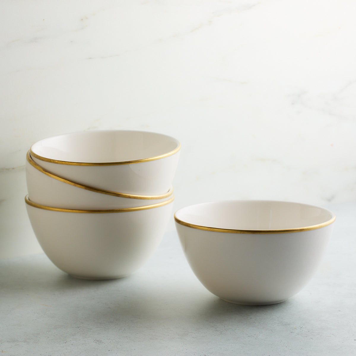 Four Caskata Artisanal Home Grace Gold Cereal Bowls, three stacked and one separate, placed on a light-colored surface with a marble background.