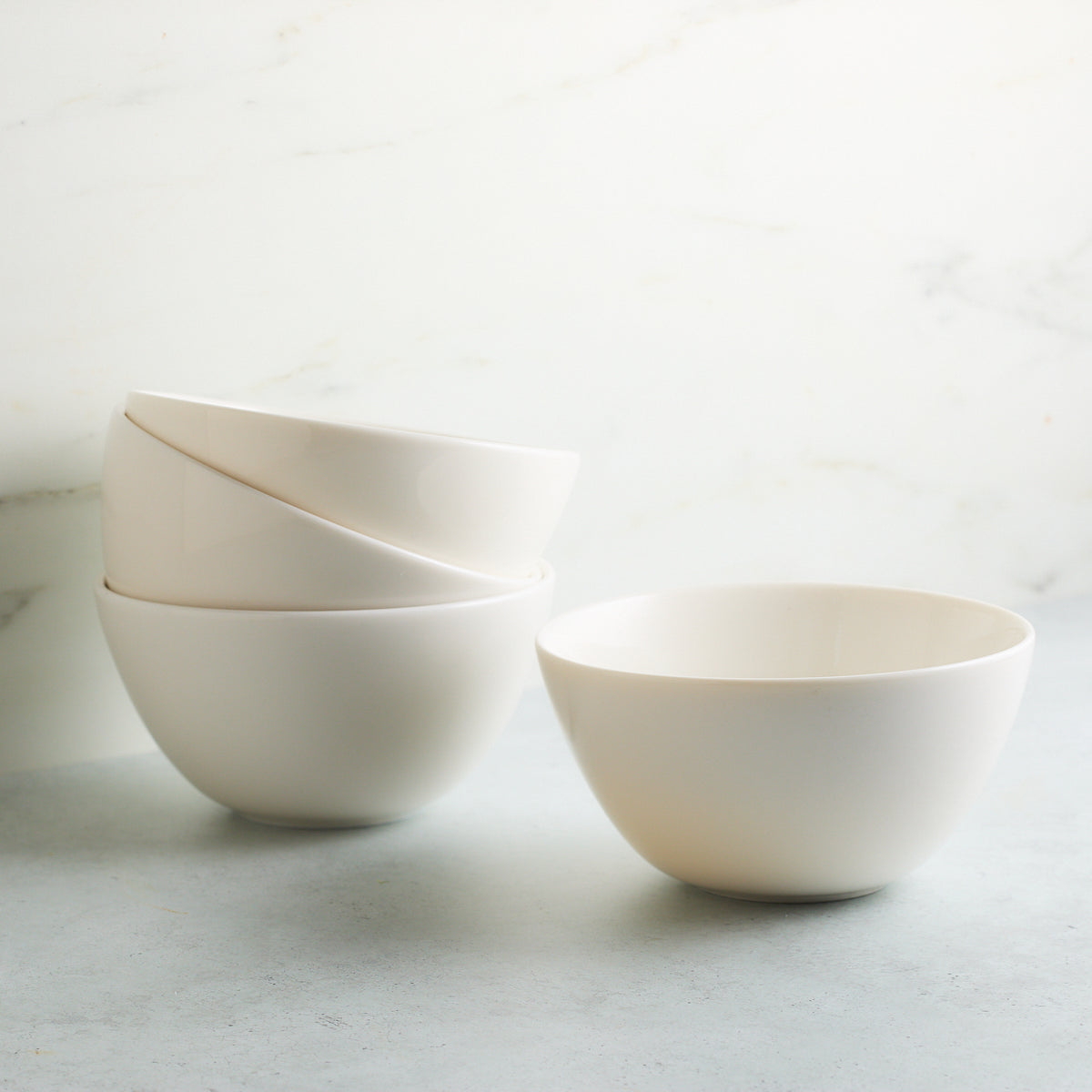 Four sleek white cereal bowls on a gray surface, with three stacked on the left and one separate on the right against a white marble background. The Grace White Cereal Bowl by Caskata Artisanal Home, made of high-fired porcelain, adds elegance to any setting.