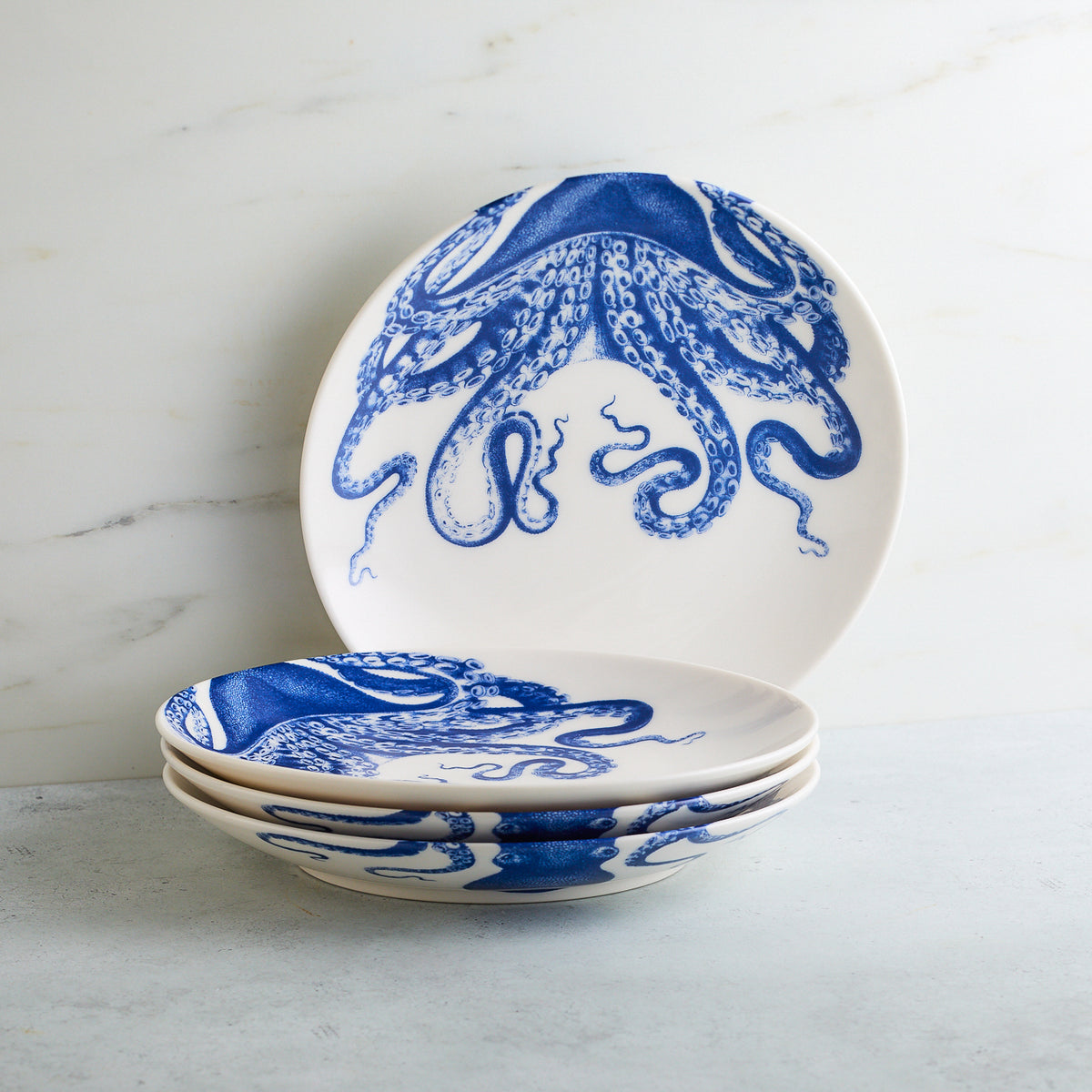 Four creamy white Lucy Coupe Salad Plates by Caskata Artisanal Home, featuring blue octopus designs, are stacked on a gray surface in front of a light-colored backdrop. These contemporary-shaped dishes offer both dishwasher and microwave safe convenience.