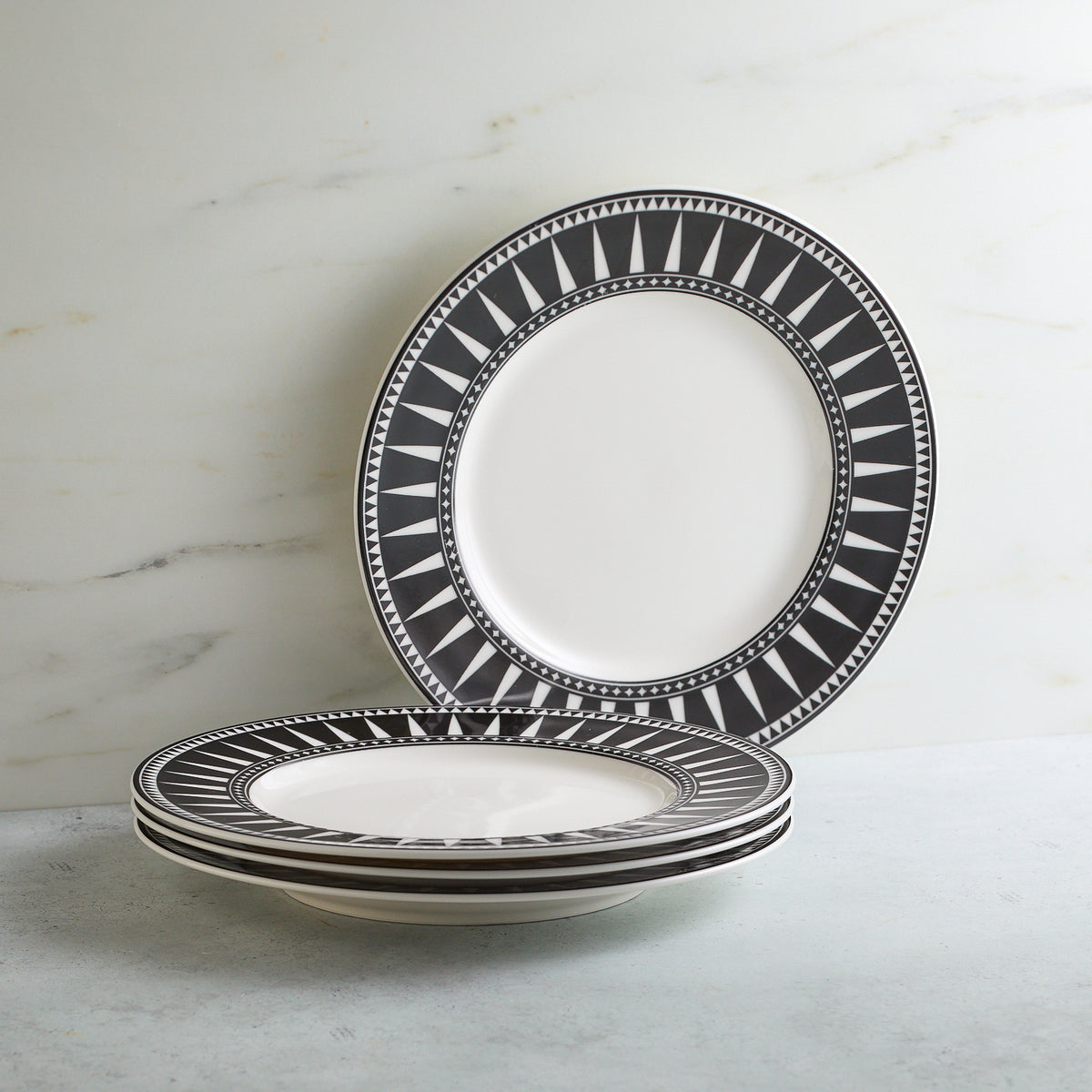 A stack of four Marrakech Rimmed Salad Plates from Caskata Artisanal Home with black and white geometric patterns along the edges, placed on a light gray surface against a white marble background.