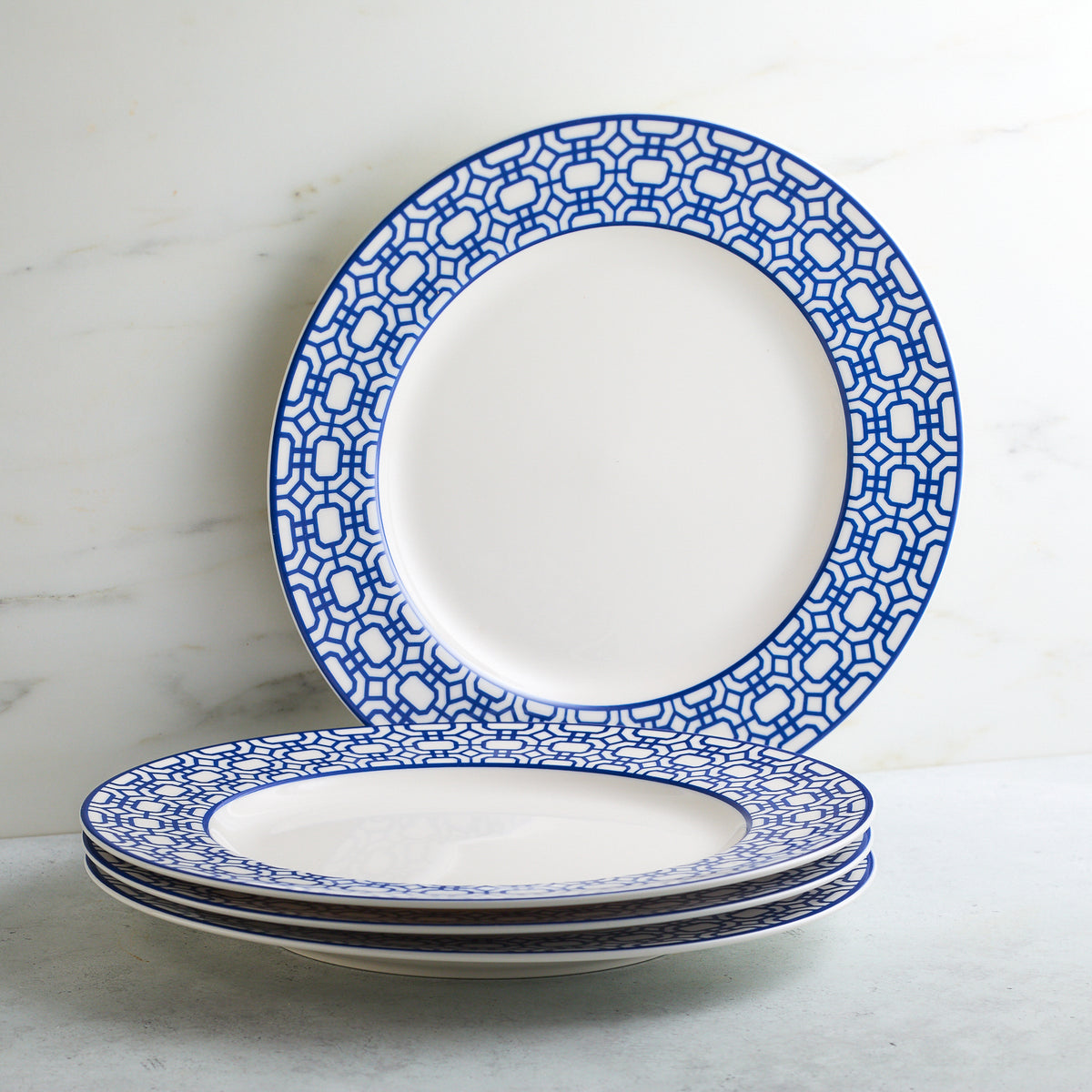 A stack of four white ceramic plates with blue geometric patterns around the edges, part of a coastal collection by Caskata Artisanal Home, sits on a light-colored surface. One Newport Garden Gate Rimmed Dinner Plate stands upright behind the stack.
