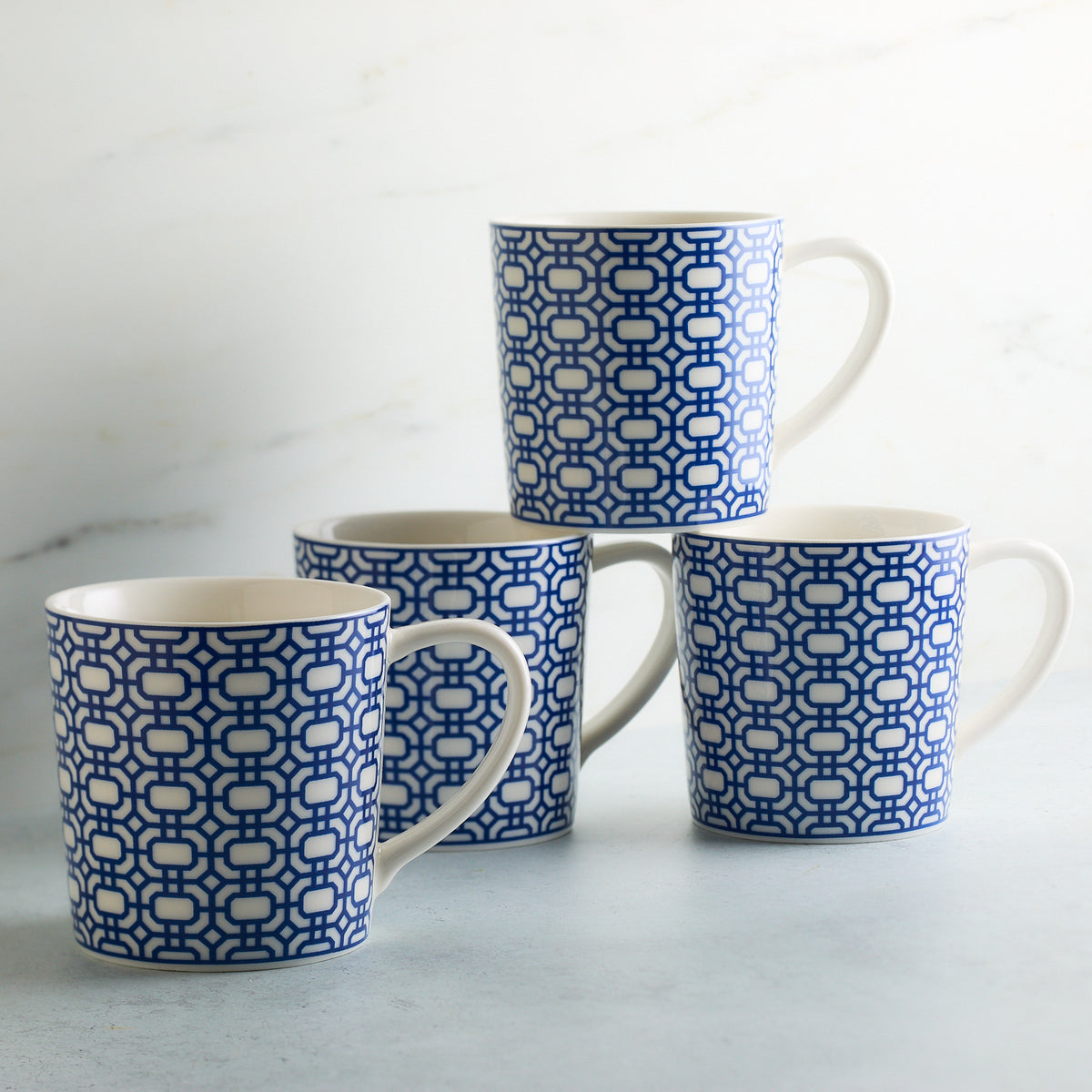 Four creamy white Newport Garden Gate Mugs by Caskata Artisanal Home with blue geometric patterns are stacked on a light gray surface against a light background. Dishwasher and microwave safe, these graphic mugs add a touch of elegance to any kitchen.