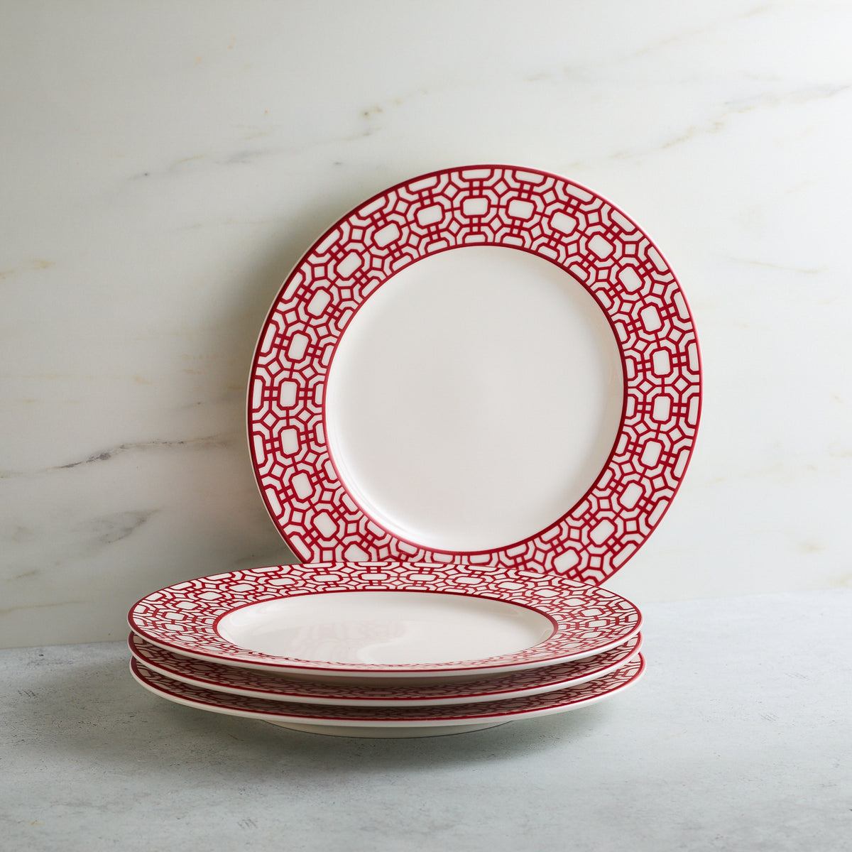 Four white ceramic salad plates with red geometric patterns are stacked on a light grey surface against a white marble background. One premium porcelain plate, the Caskata Artisanal Home Newport Garden Gate Crimson Rimmed Salad Plate, stands upright at the back.