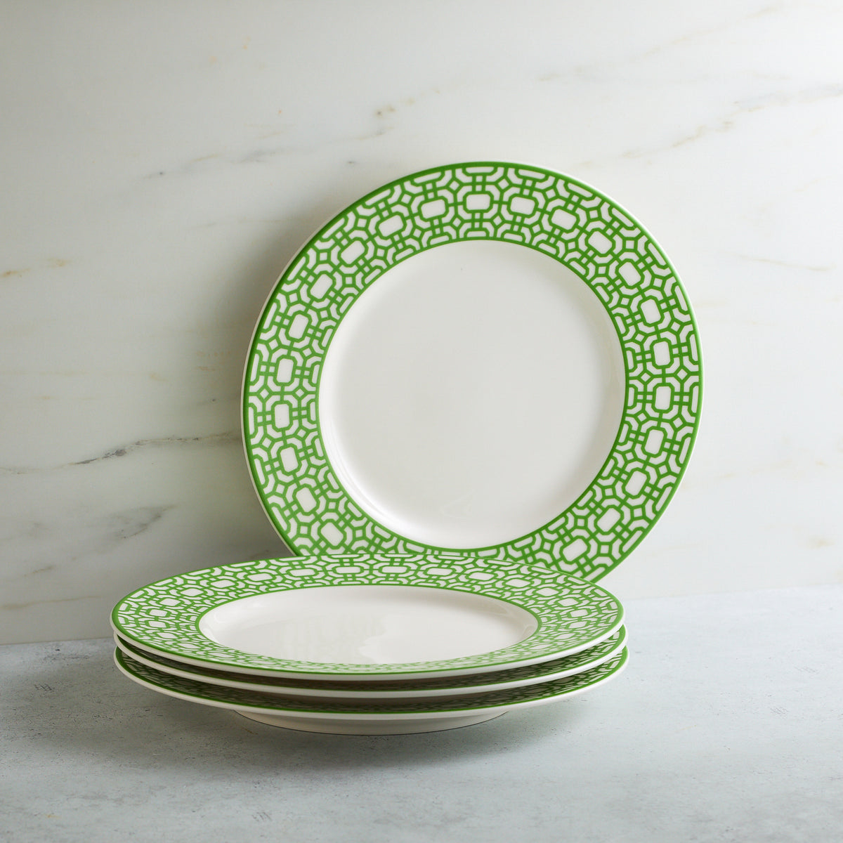 A stack of four Newport Garden Gate Verde Rimmed Salad Plate by Caskata Artisanal Home, placed on a light gray surface against a marble background. These porcelain salad plates add a touch of contemporary tableware elegance to any setting.
