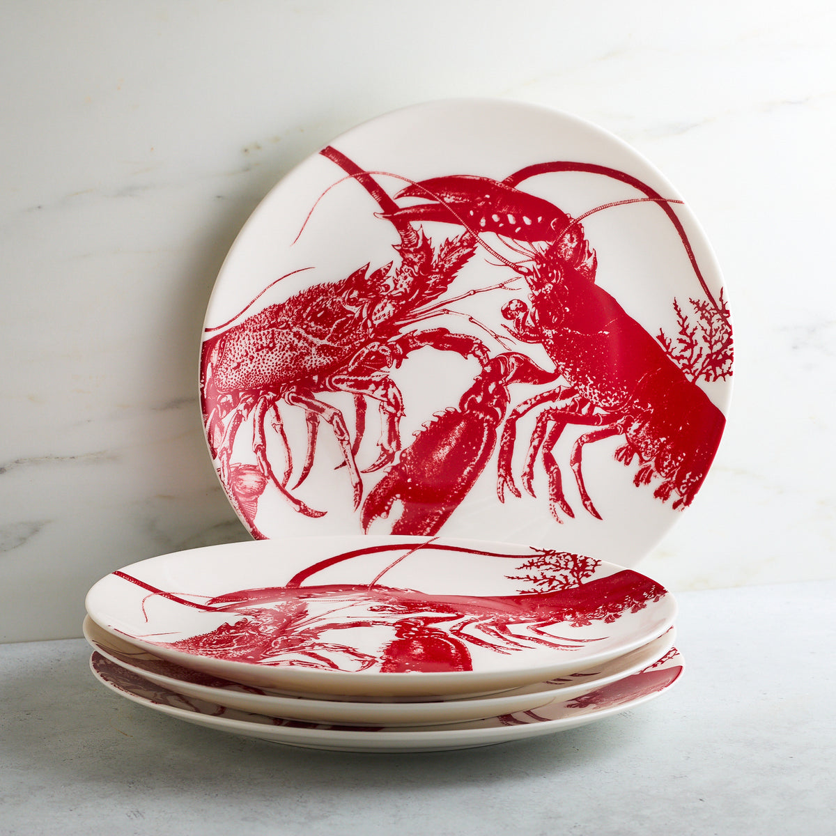 A set of premium porcelain plates with red lobster designs, stacked neatly on a white marble surface. Dishwasher and microwave safe, these seaside style Lobster Coupe Dinner Plates by Caskata add a touch of coastal charm to your dining experience.