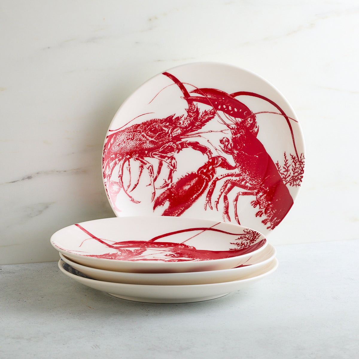 Four Lobster Coupe Salad Plates by Caskata featuring red lobster illustrations are stacked on a light gray surface. One plate is upright, leaning against a white marble backdrop. These premium porcelain plates bring a touch of seaside style to your dining experience.