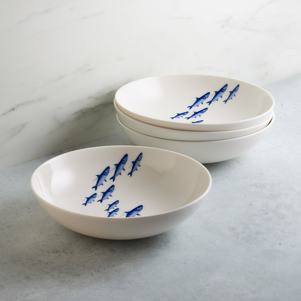 Three white ceramic bowls with blue, school of fish designs are stacked on a gray countertop near a marble wall. Two additional bowls with the same design are in the foreground, all part of the charming Caskata Artisanal Home School Fish Entrée Bowl collection.