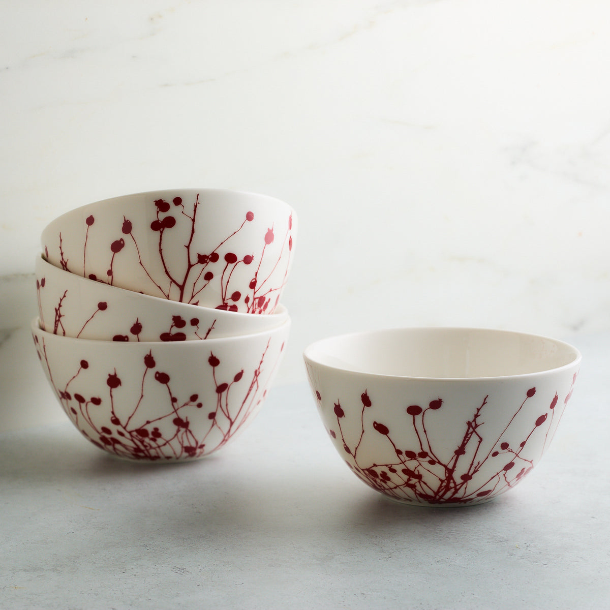 Four Winterberries Cereal Bowls by Caskata Artisanal Home, adorned with red floral patterns reminiscent of winter berries, are arranged on a light grey surface against a white background. Three bowls are stacked neatly on the left, while one stands alone on the right. These dishwasher-safe bowls add a touch of elegance to any table setting.