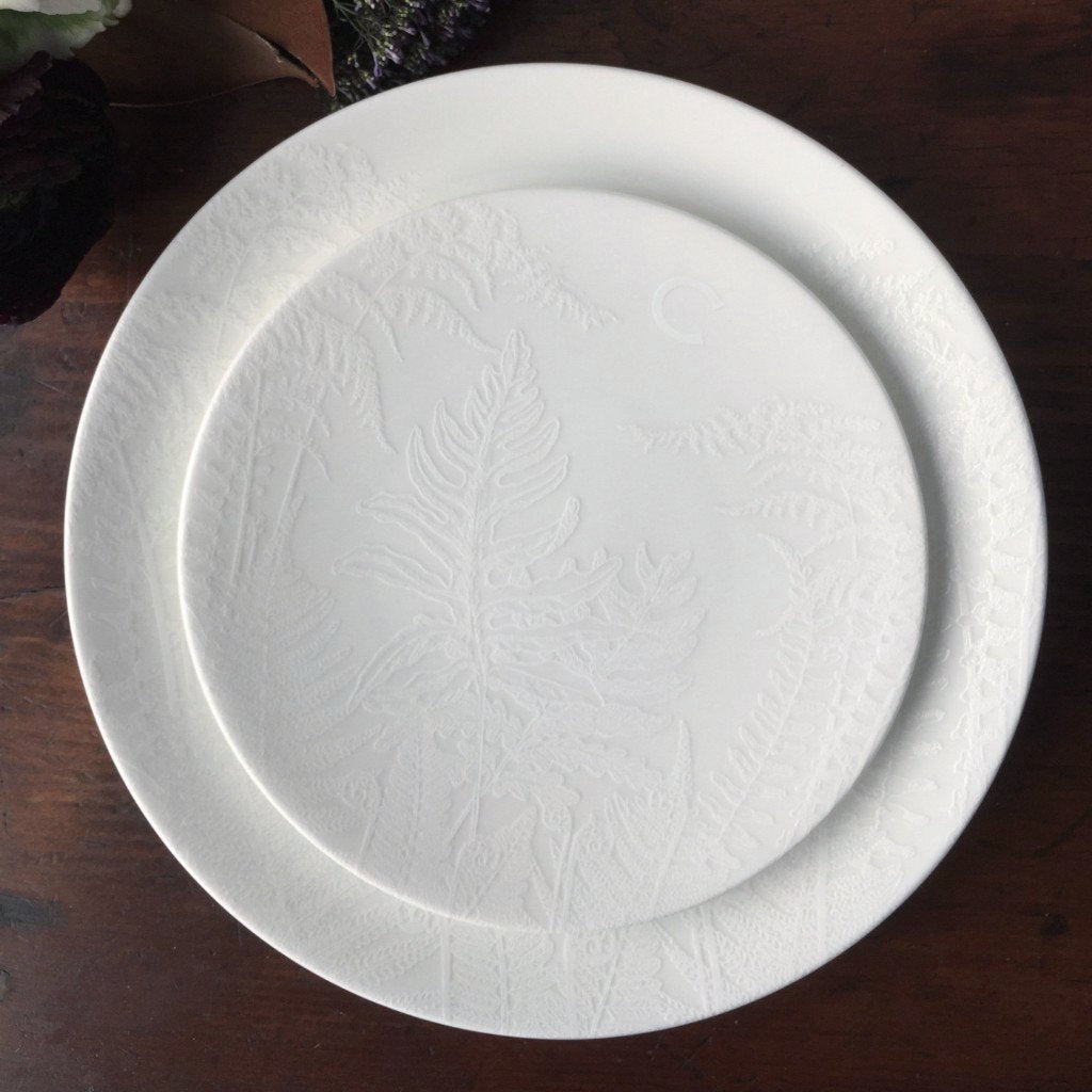 Two Spring Small Plates with embossed fern patterns, part of the Caskata Artisanal Home collection. The larger plate is underneath the smaller one, and both rest elegantly on a dark wooden surface.