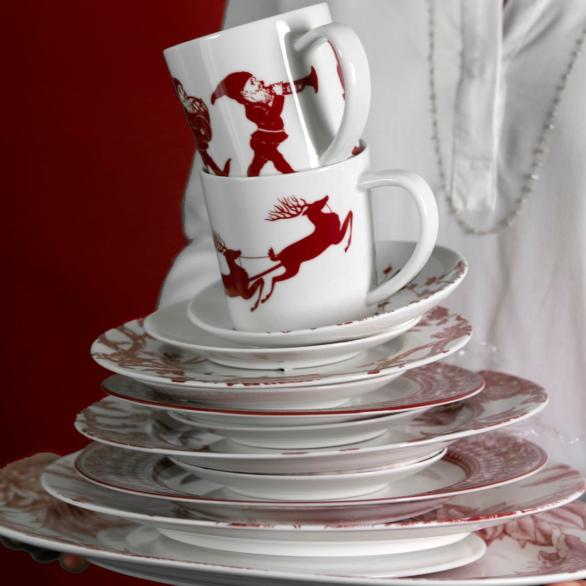 A stack of Elves Mugs with reindeer and Santa Claus designs, part of a festive holiday collection by Caskata Artisanal Home, held by a person in a white garment.