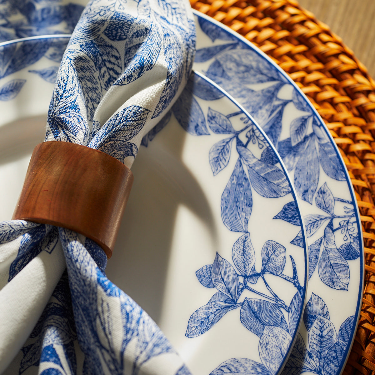 Close-up of a table setting with heirloom-quality dinnerware featuring Caskata Artisanal Home Arbor Rimmed Dinner Plate blue floral-patterned plates, a napkin with a similar design held by a wooden ring, and a woven placemat beneath.