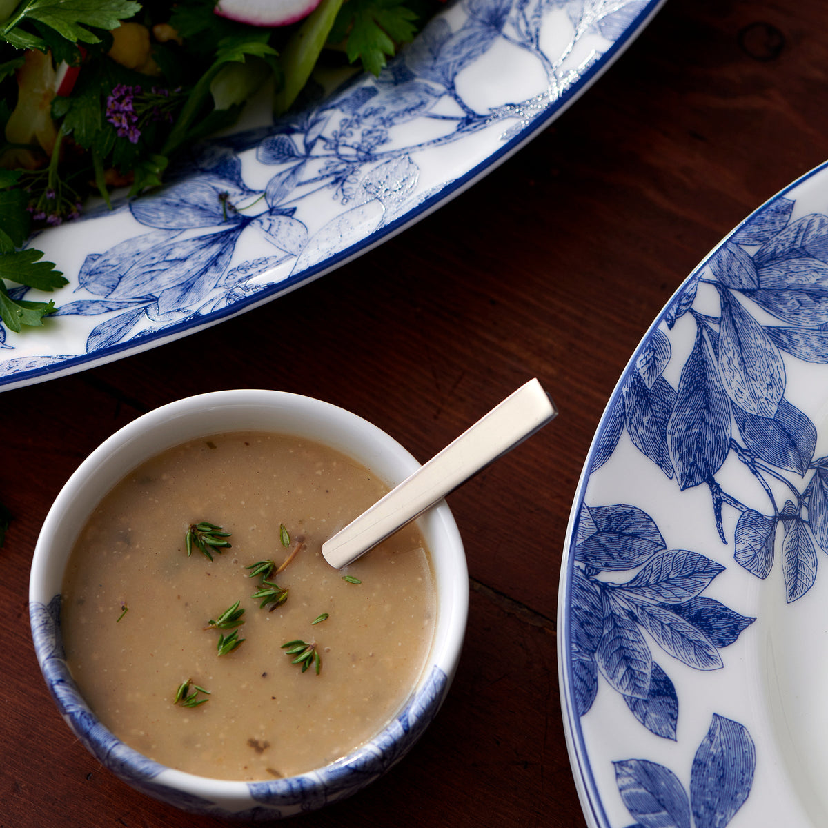 A small bowl of creamy soup garnished with herbs, placed next to a Caskata Artisanal Home Arbor Snack Bowl adorned with blue floral patterns and leafy branches on a wooden table.