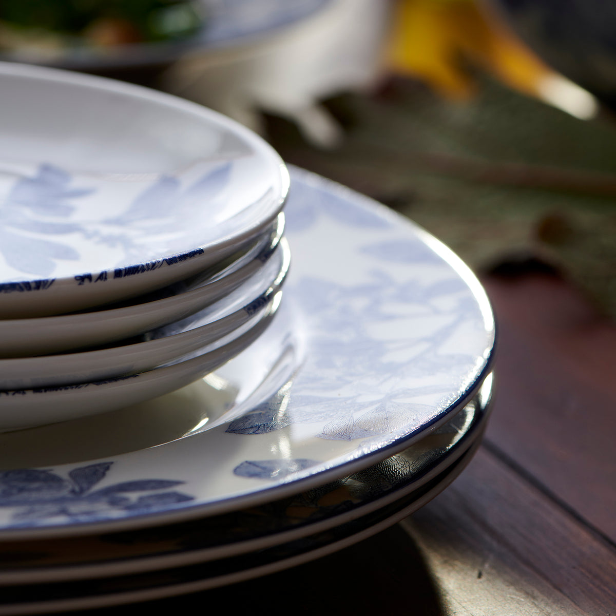 Close-up of a stack of Arbor Blue Small Plates by Caskata Artisanal Home with blue floral patterns on a wooden table. Sunlight casts shadows on the premium porcelain plates, highlighting their intricate botanical details.
