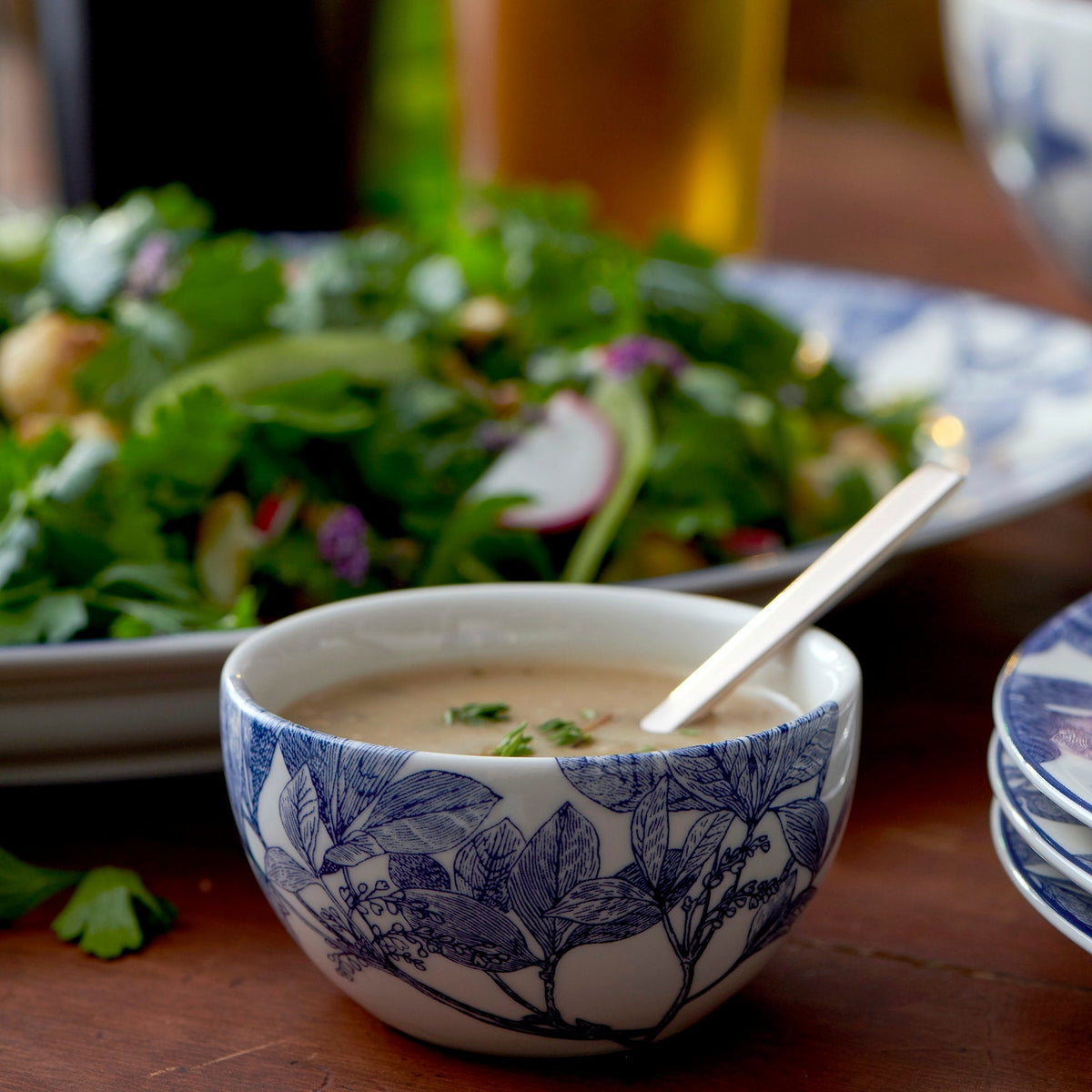 A dishwasher and microwave safe ceramic bowl with a blue floral pattern contains a spoon resting in a light-colored soup or sauce. In the background, there is a mixed salad with greens and radishes on a white plate. The bowl is an Arbor Snack Bowl from Caskata Artisanal Home.