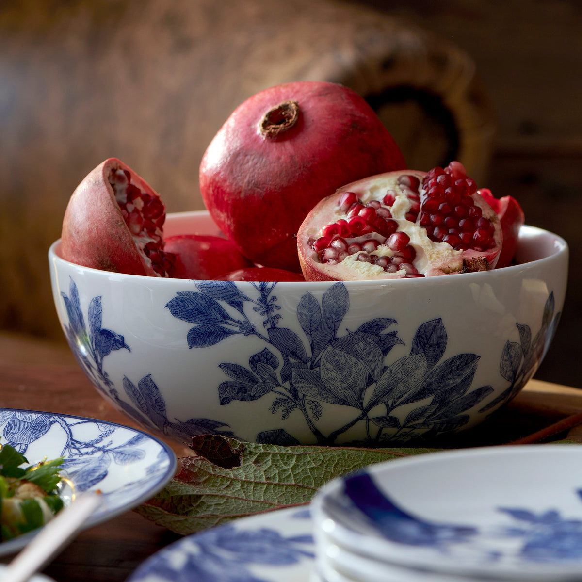 A premium porcelain Arbor Vegetable Serving Bowl from Caskata Artisanal Home with blue floral patterns holds whole and halved pomegranates, placed on a dining table with similarly patterned plates. This dishwasher-safe piece adds elegance and convenience to your dining experience.