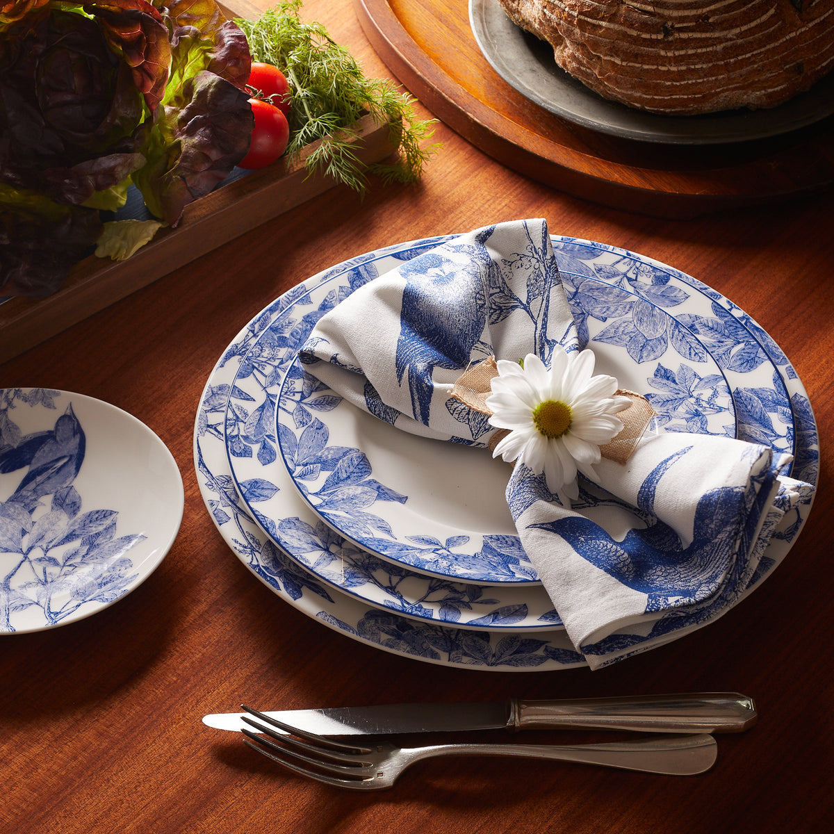 A table setting with blue and white floral-patterned Arbor Rimmed Dinner Plates from Caskata Artisanal Home, showcasing heirloom-quality dinnerware, is stacked with a napkin and daisy on top. Silverware lies beside, and vegetables are in the background.