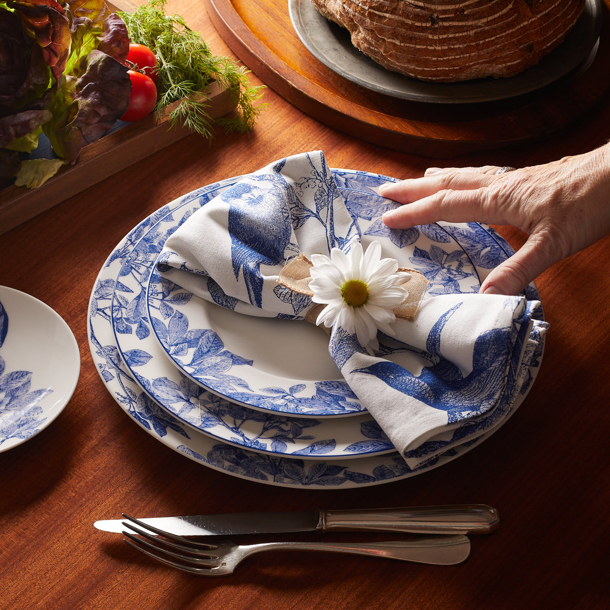 A hand adjusts a napkin with a daisy on an Arbor Rimmed Charger Plate by Caskata Artisanal Home, placed on a wooden table with cutlery, bread, and vegetables in the background.