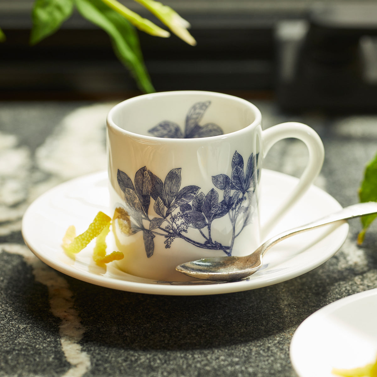 A white ceramic cup with blue floral patterns on a matching saucer, crafted from bone china, with a silver spoon and lemon peel beside it, the Arbor Espresso Cups &amp; Saucers, Set of 2 by Caskata.