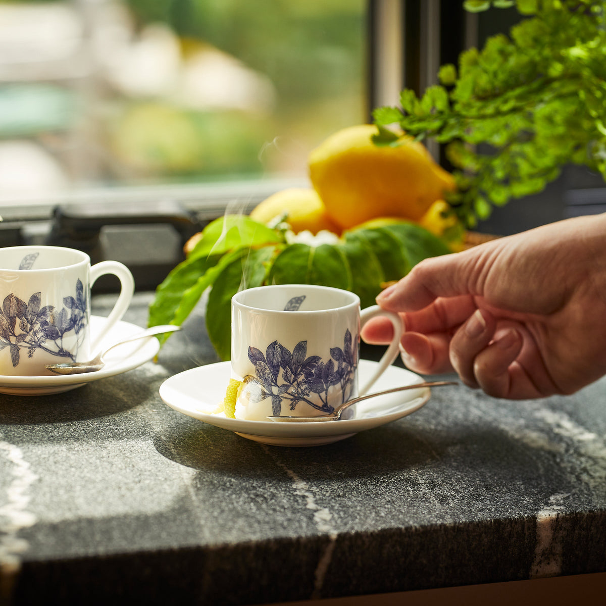 A hand reaching for a teacup with a saucer and spoon, beside another cup on a countertop. Two lemons and a green plant are in the background near a window, highlighting the elegance of Arbor Espresso Cups &amp; Saucers, Set of 2 by Caskata.