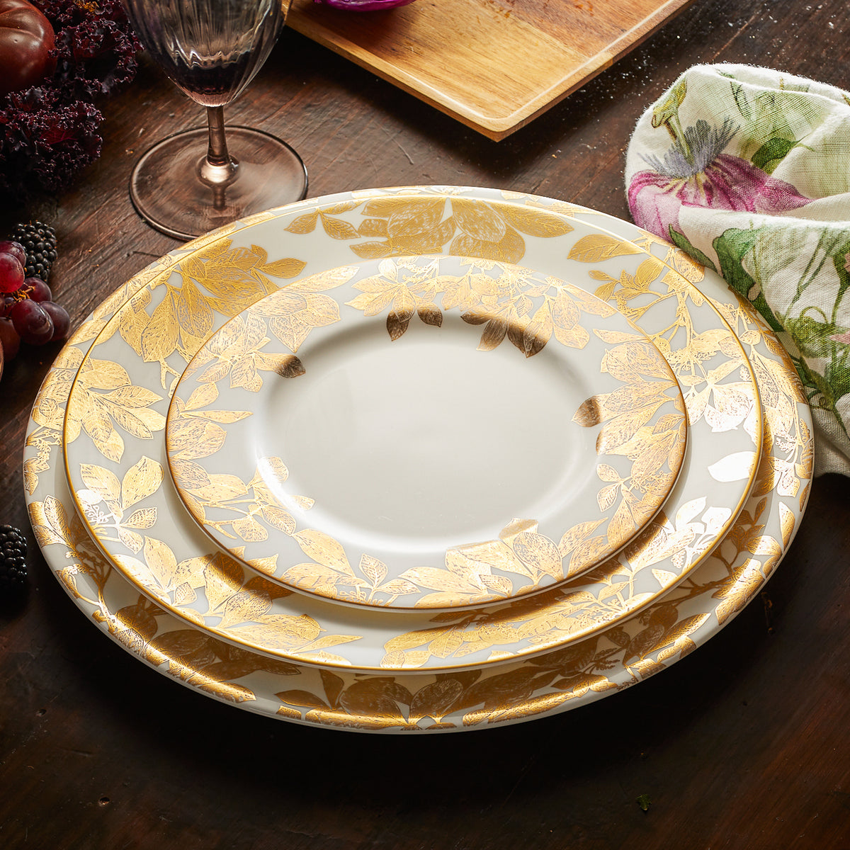 Three stacked Arbor Gold Rimmed Charger plates by Caskata Artisanal Home, adorned with intricate gold leaf patterns, are displayed on a wooden table, accompanied by a wine glass, a patterned napkin, and assorted fruits in the background.