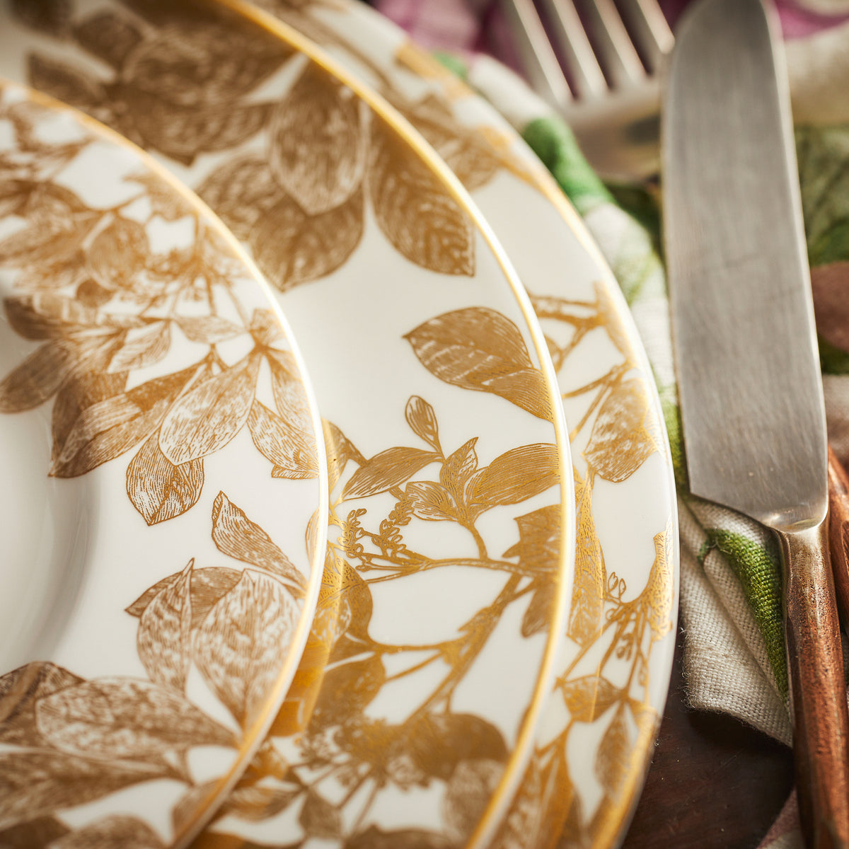 Close-up of three stacked Caskata Artisanal Home Arbor Gold Rimmed Chargers with a golden leaf pattern, next to a knife and a fork.