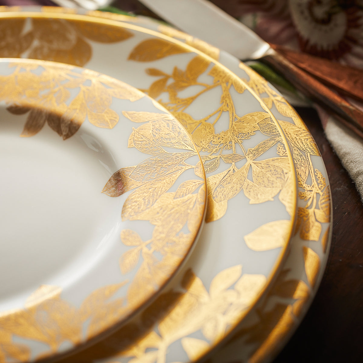 Close-up of three overlapping Arbor Gold Rimmed Charger plates by Caskata Artisanal Home with intricate gold leaf and botanical details on their rims, placed on a wooden surface.