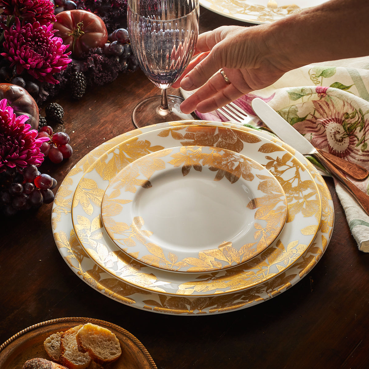 A hand arranges a goblet on a table set with Caskata Artisanal Home&#39;s Arbor Gold Rimmed Charger, surrounded by botanical details, flowers, fruit, and bread.