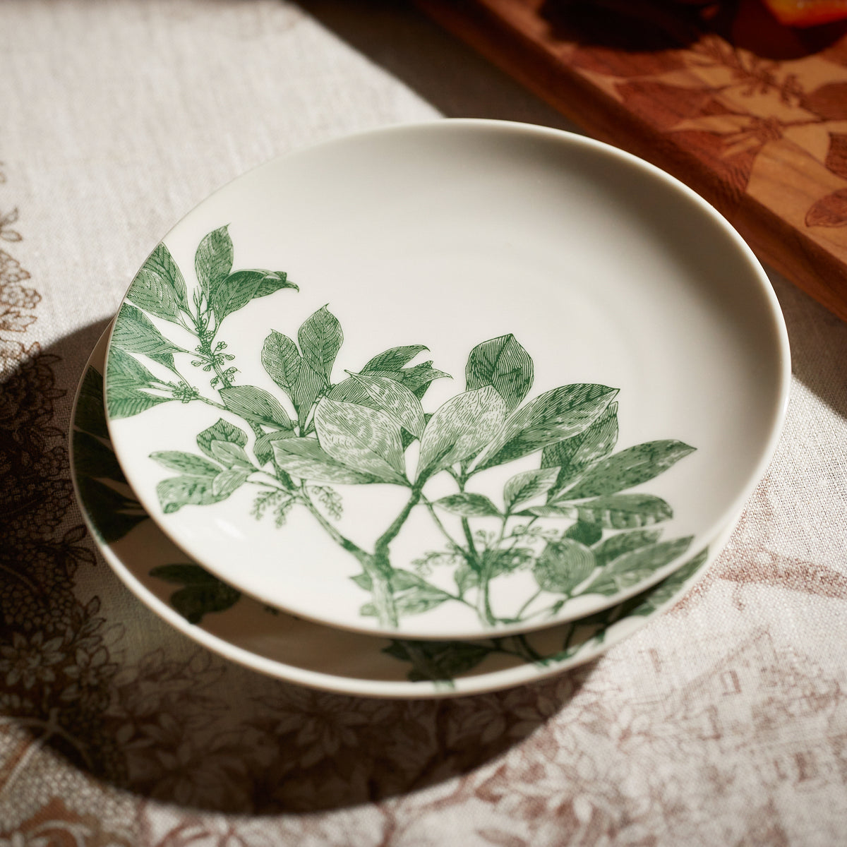 A white ceramic plate with green leaf designs, part of a botanical dinnerware collection, sits on a patterned tablecloth, partially overlapping another plate beneath it. This elegant Arbor Green Small Plate by Caskata is also conveniently dishwasher safe.