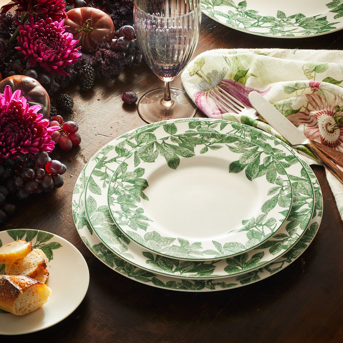 A table is set with Caskata Arbor Green Rimmed Dinner Plate, accompanied by a floral-patterned napkin, bread on a side plate, a glass, and a centerpiece of flowers and fruits.