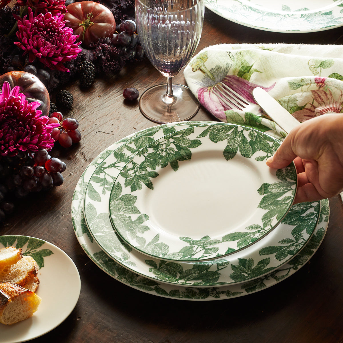 A hand setting a Caskata Arbor Green Rimmed Salad Plate with green leaf patterns on a table, next to larger plates, a drinking glass, a floral napkin, purple flowers, grapes, and a small bread plate.