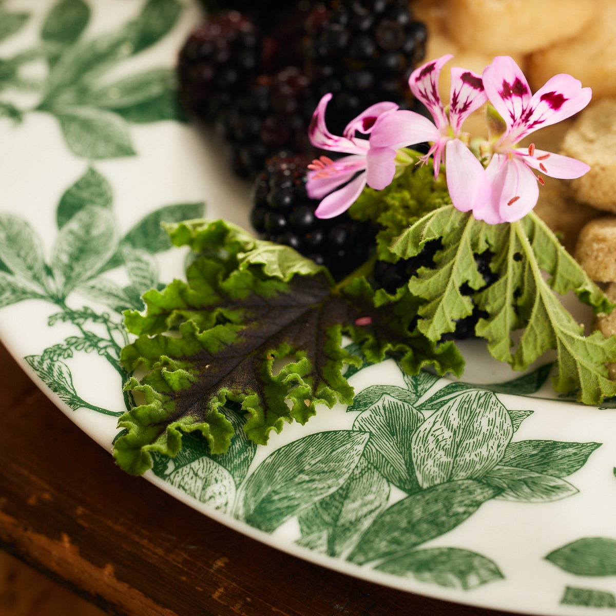 Close-up of a decorative Caskata Arbor Green Rimmed Dinner Plate made of premium porcelain, featuring botanical details with green leafy designs. It holds a small arrangement of fresh blackberries, a pink flower, and leafy greens.