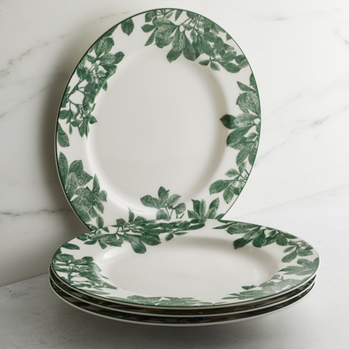 A stack of three premium Caskata Arbor Green Rimmed Dinner Plates, adorned with green foliage patterns along the rim, with one additional plate showcasing its botanical details resting upright behind the stack.