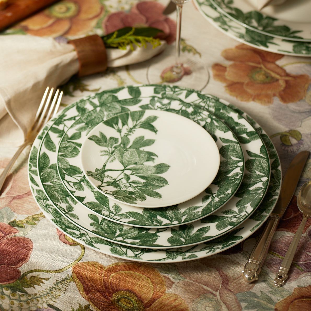 A set of Arbor Green Rimmed Salad Plates by Caskata with green leaf patterns stacked on a floral tablecloth. Silverware and a napkin with a wooden ring are placed beside the salad plates.