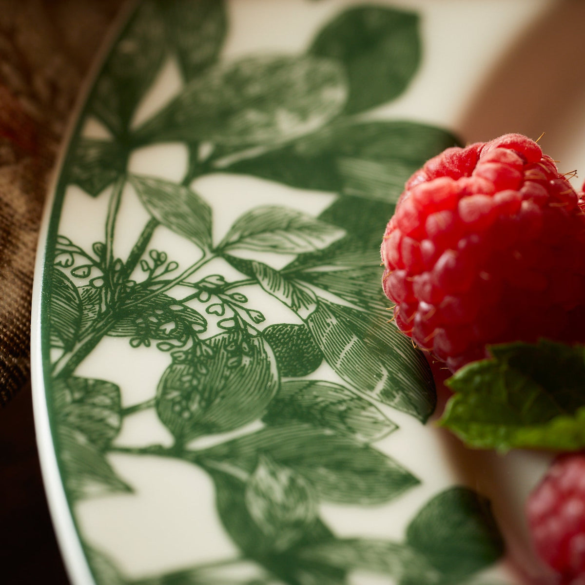 Close-up of a premium porcelain Arbor Green Rimmed Salad Plate by Caskata adorned with green foliage designs, featuring fresh raspberries and mint leaves on the edge.