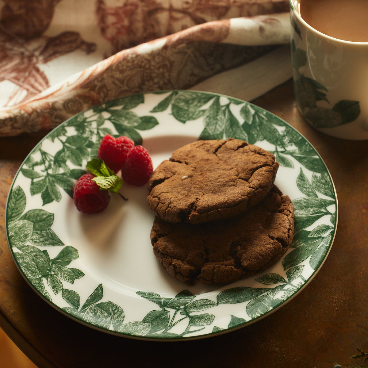 A Caskata Arbor Green Rimmed Salad Plate made from premium porcelain with a green leaf pattern holds two chocolate cookies and a few raspberries, accompanied by a cup of beverage.