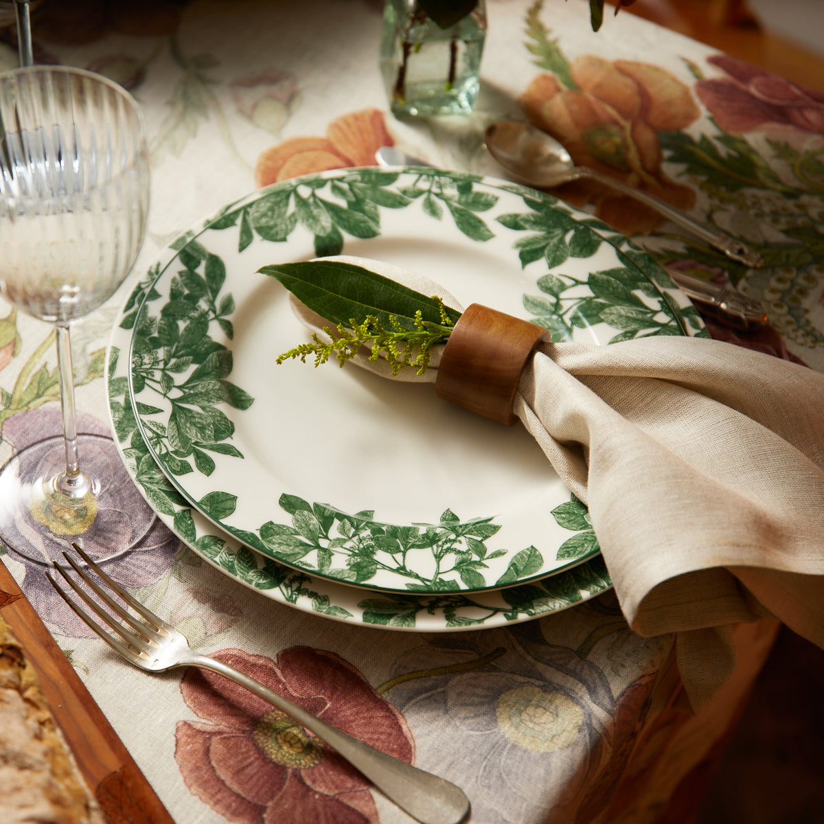Close-up of a table setting featuring a floral tablecloth, an Arbor Green Rimmed Dinner Plate by Caskata, a cloth napkin with a wooden napkin ring, silver cutlery, and a wine glass.